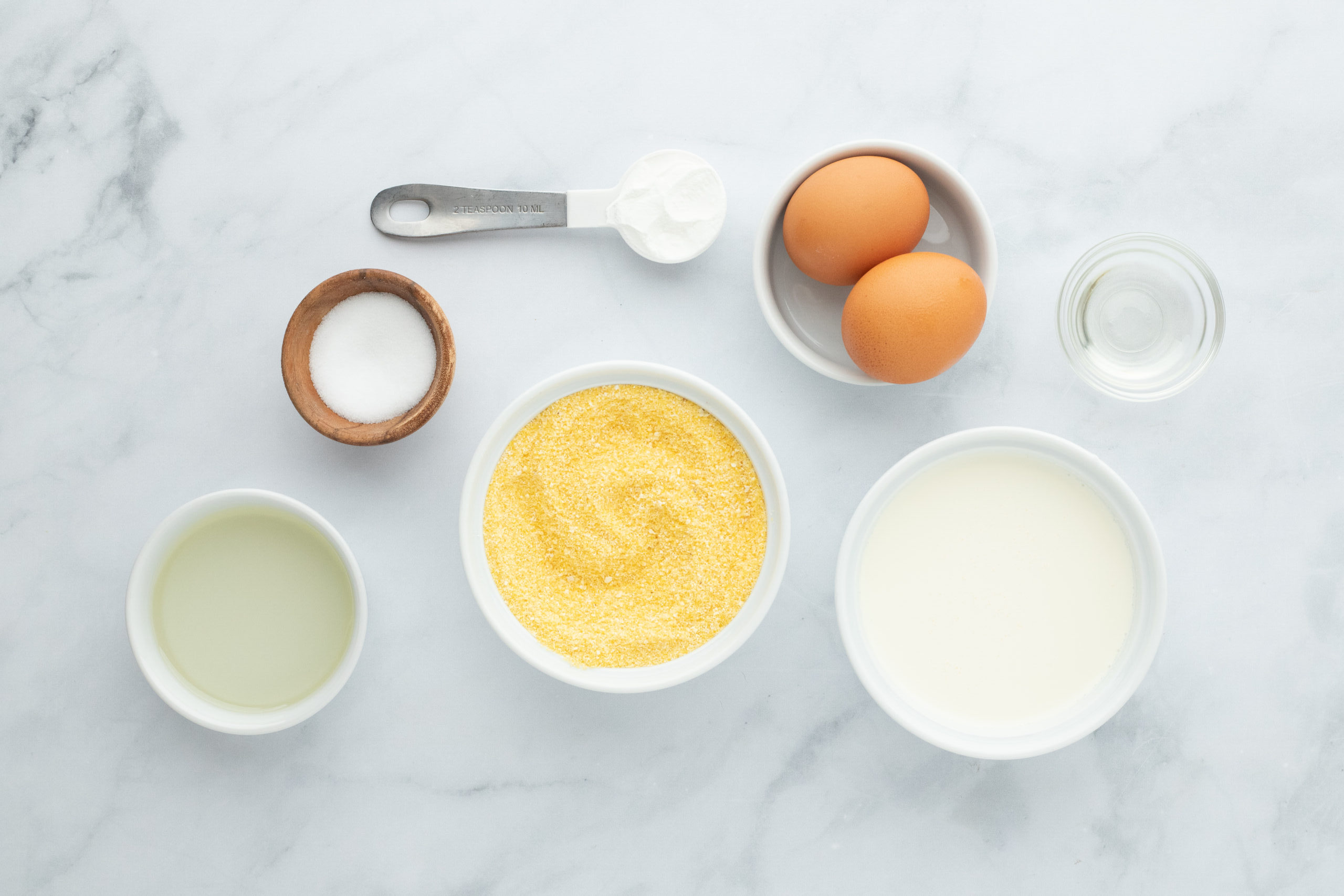 Cornmeal, heavy cream, bacon fat, salt, eggs, hot water in white bowls on a white background