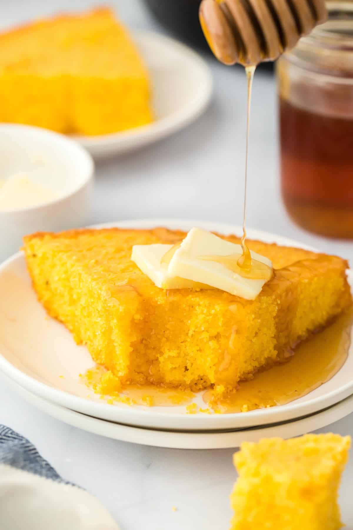 A slice of Southern corn pone on a white plate with butter and honey being drizzled on top on a white background