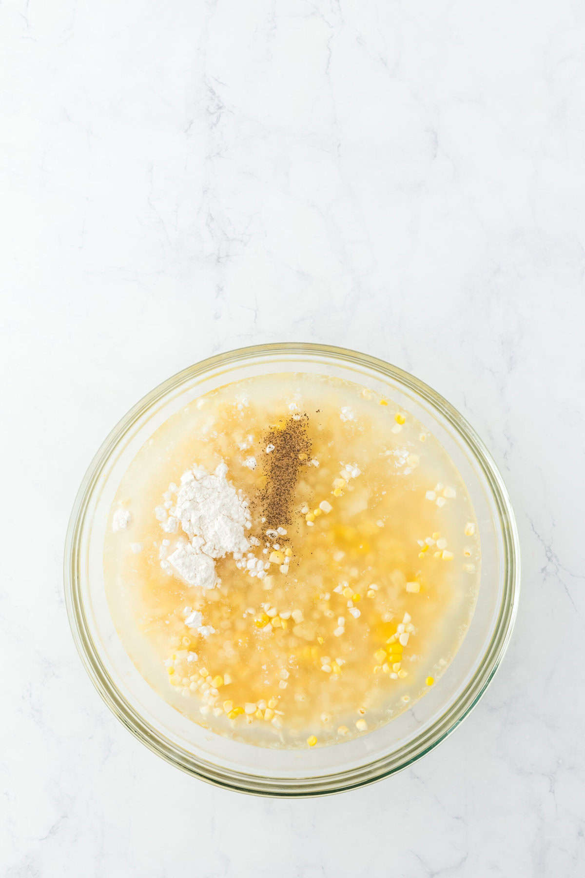 Flour, sugar, salt and pepper added to fresh corn kernels in a glass bowl with water in a white background