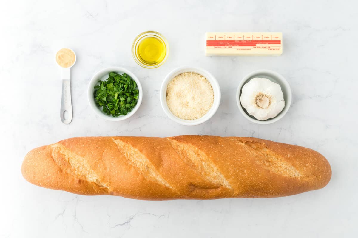 Butter, a baguette, garlic, parsley, parmesan, garlic powder, and olive oil on a white background