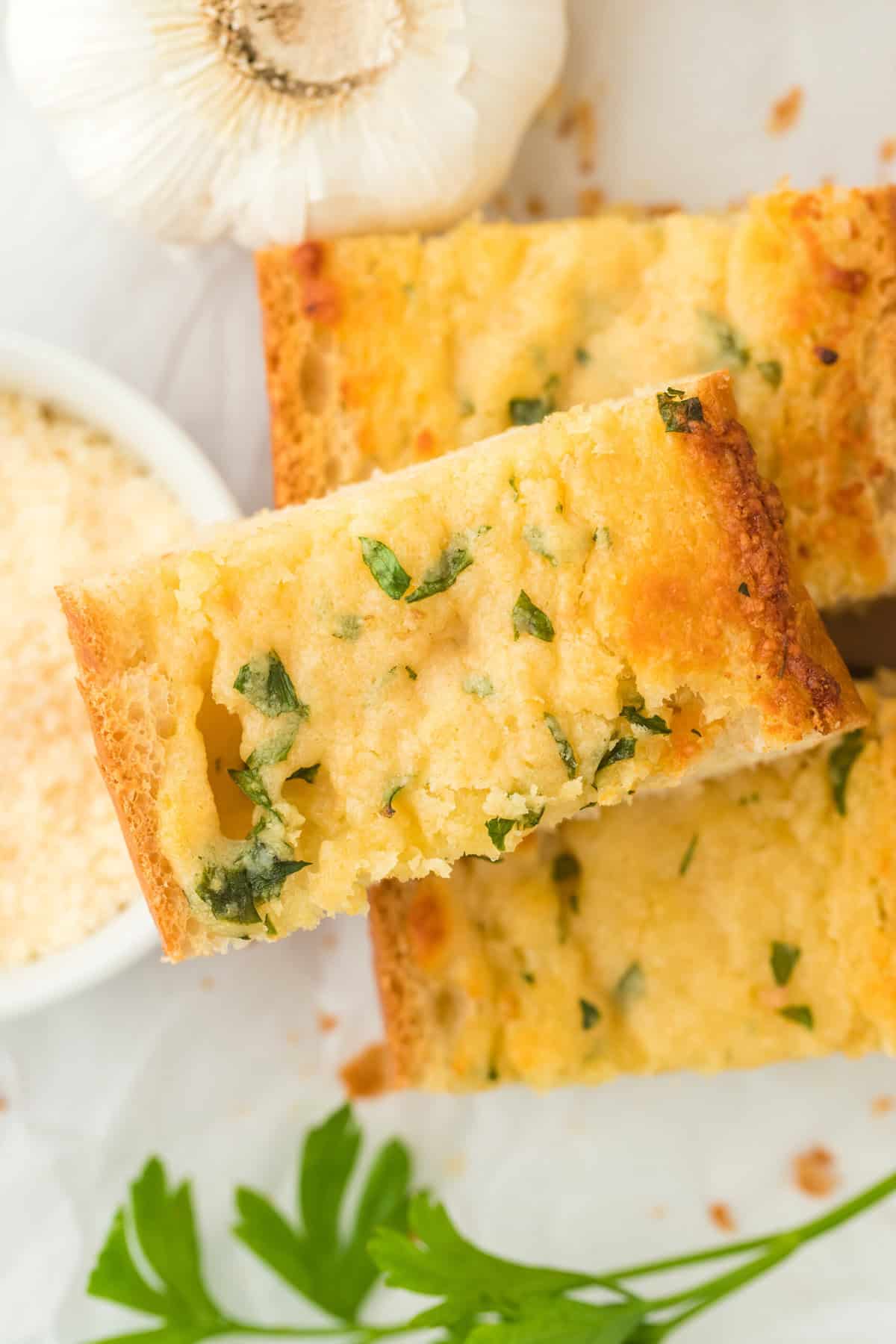 Overhead shot of the inside of garlic bread from scratch on a white plate with parsley in front of it, a head of garlic behind and a bowl with parmesan cheese next to it