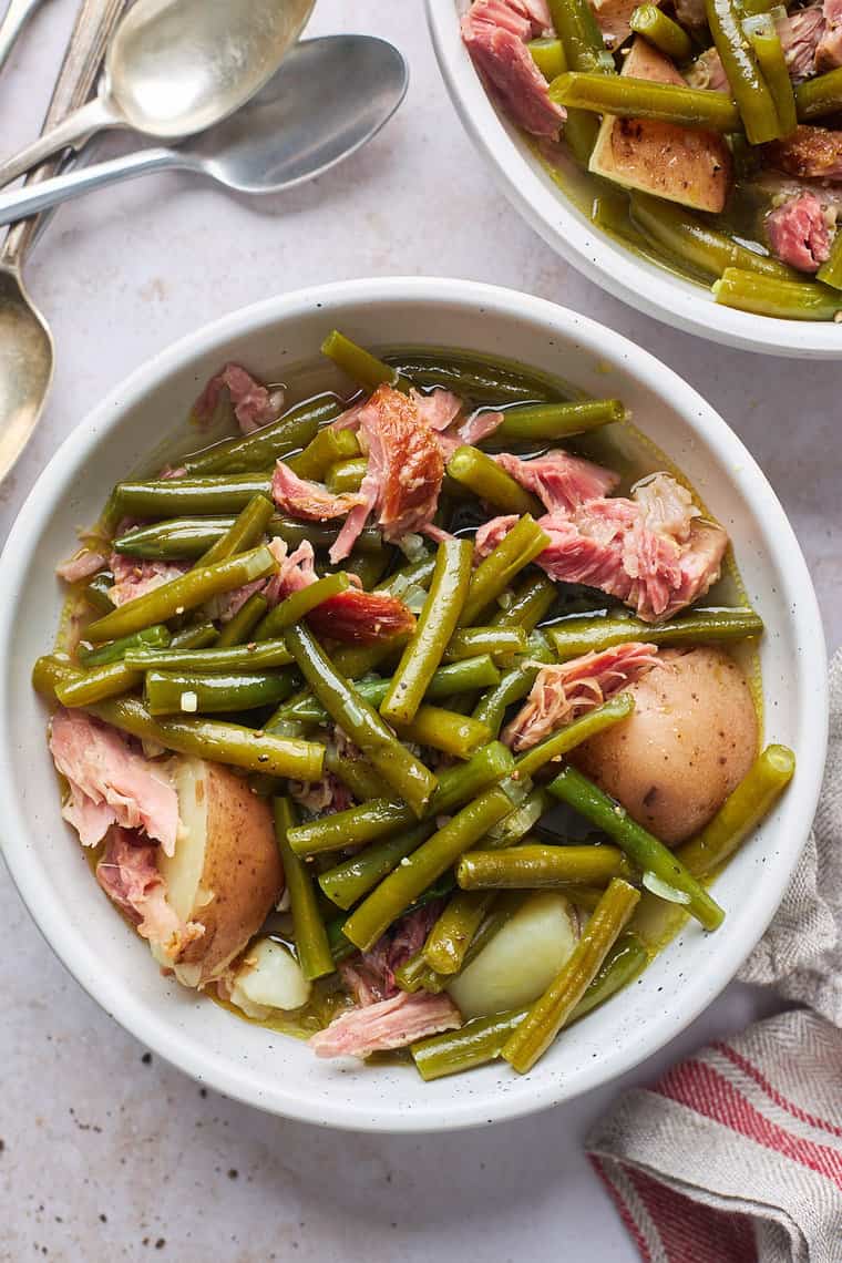 Two bowls of Southern style green beans with potatoes in them with ham hock on a white background