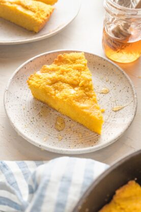 A slice of corn pone on a white speckled plate with honey and butter drizzled on top