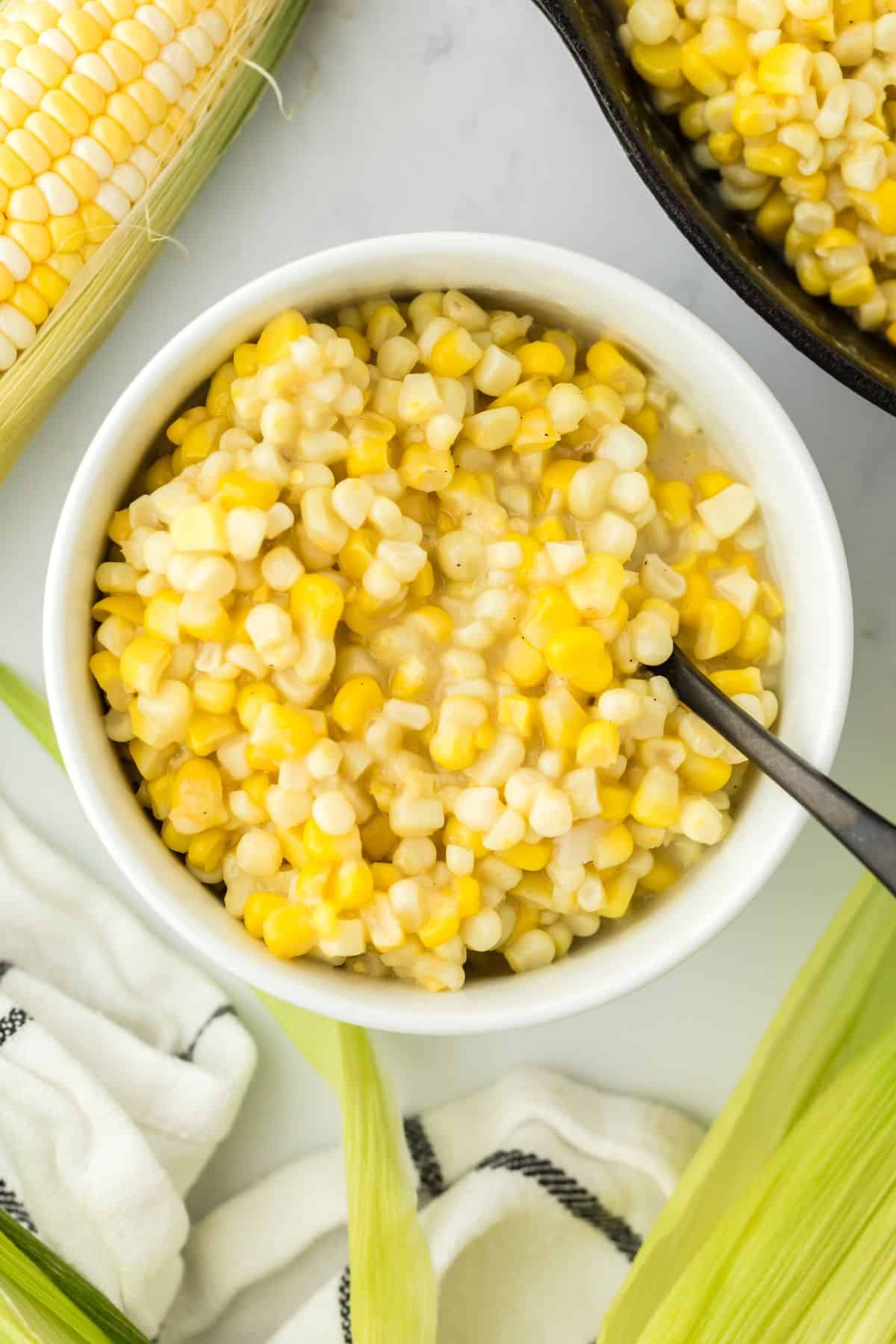 A bowl of fried corn recipe on the table with ears of fresh corn and a spoon in the bowl.