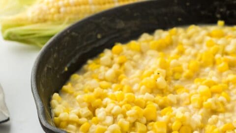 A skillet of fried corn only half shown in the image with an ear of fresh corn in the background.