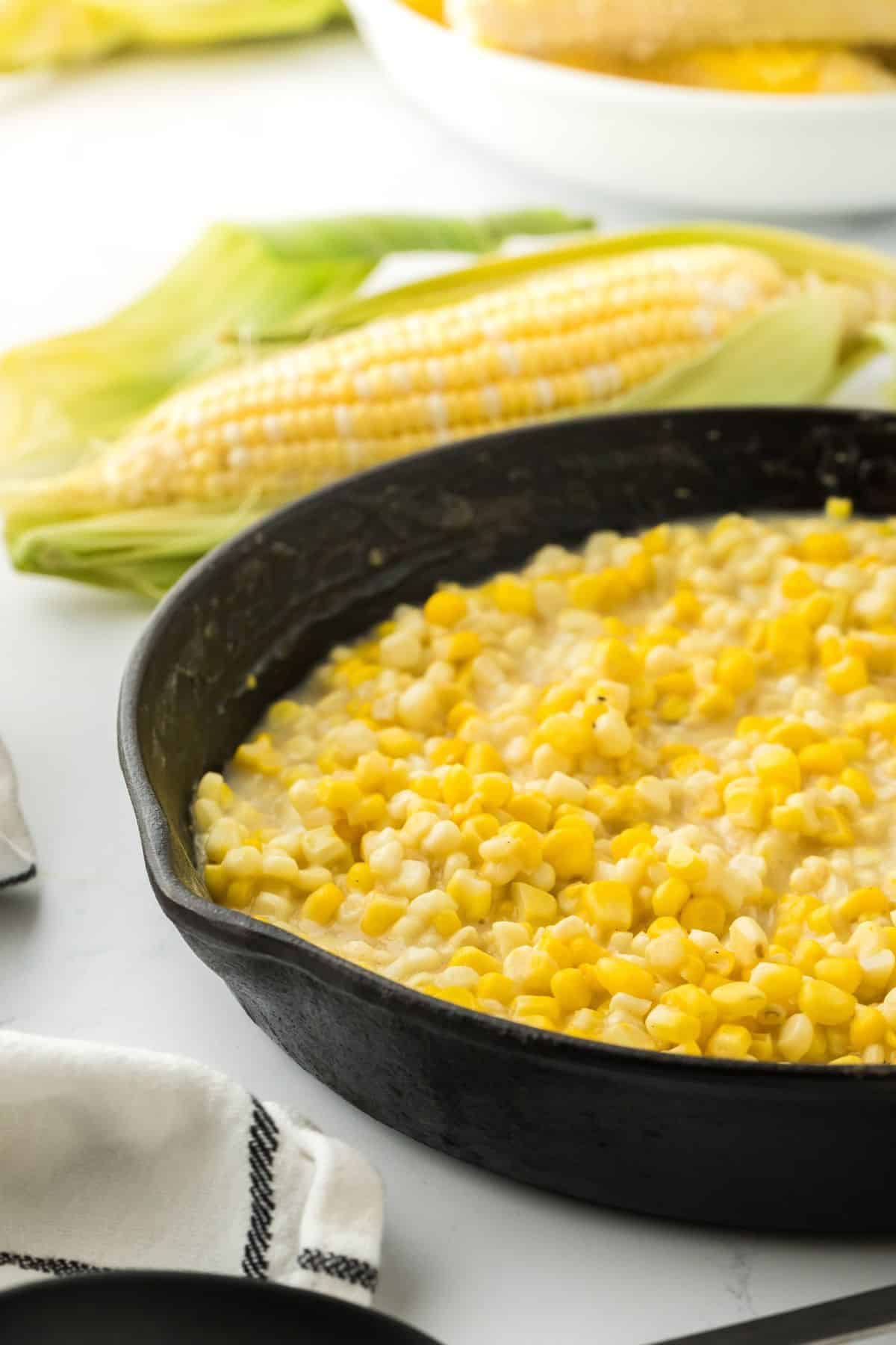 A cast iron skillet of Southern fried corn only half shown in the image with an ear of fresh corn in the background.