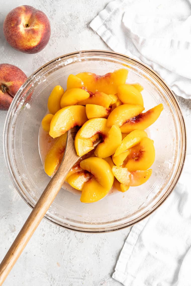 Peach slices being stirred with a wooden spoon in a glass bowl