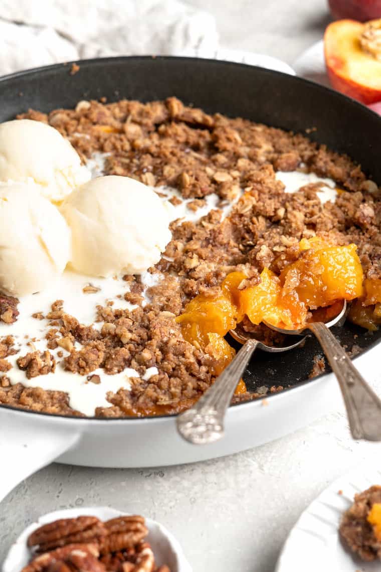 A close up of a homemade peach crisp recipe with two spoons digging in with ice cream melting on top