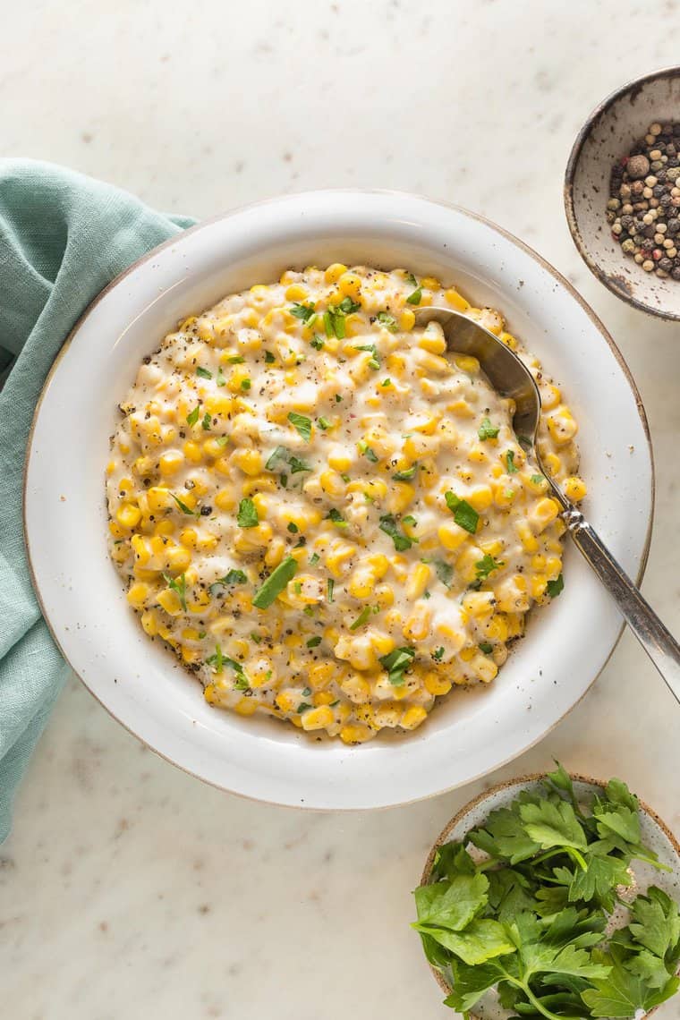 A large white bowl filled with creamed corn recipe ready to serve with a spoon inside of it