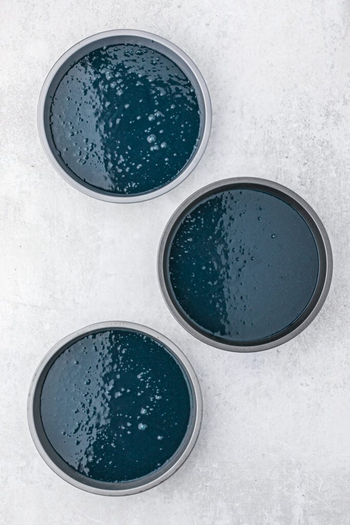 Three cake pans filled with blue tinted cake batter before baking