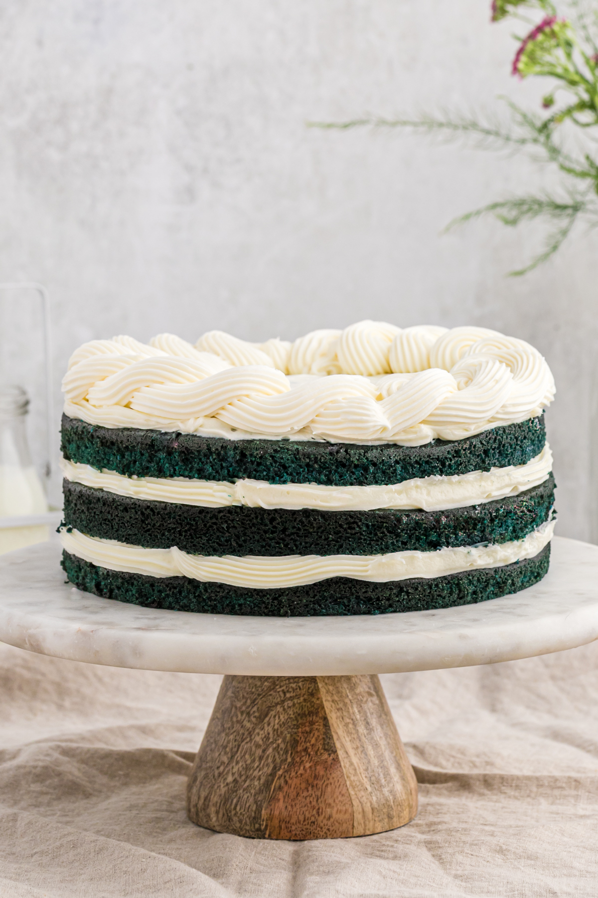 A blue velvet cake recipe with cream cheese frosting on a white cake stand with a wooden bottom against a white background