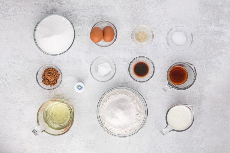 Cake ingredients like eggs, flour, oil, cocoa powder and vanilla in glass bowls on white countertop