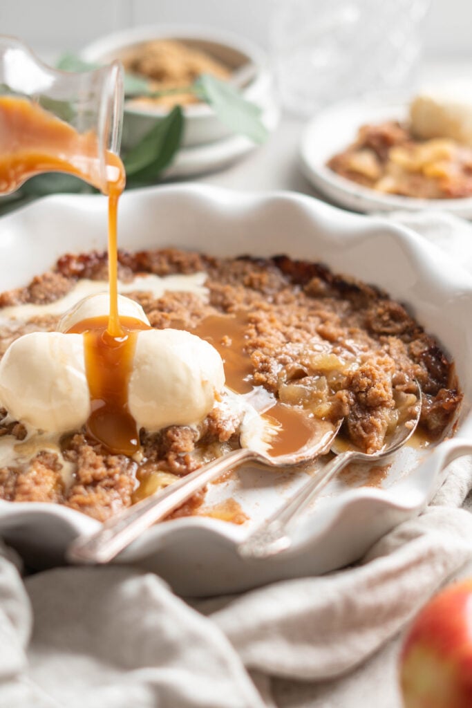 Caramel sauce being poured over a fresh apple brown betty recipe with vanilla ice cream on top