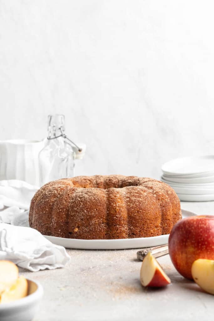 An apple cider doughnut cake recipe on a white plate against a white background with a whole apple