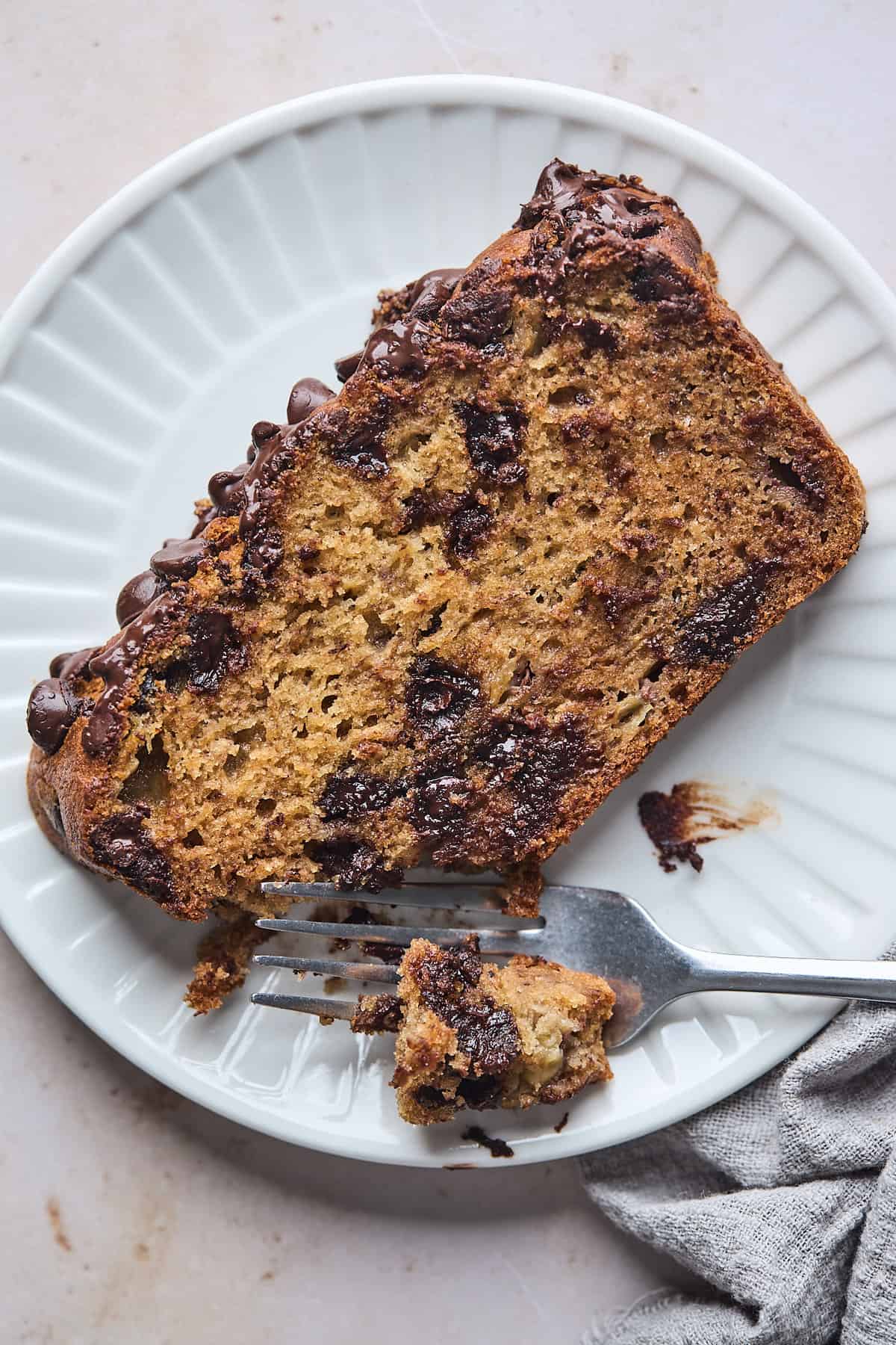 A slice of chocolate chip banana bread being eaten with a fork on a white plate