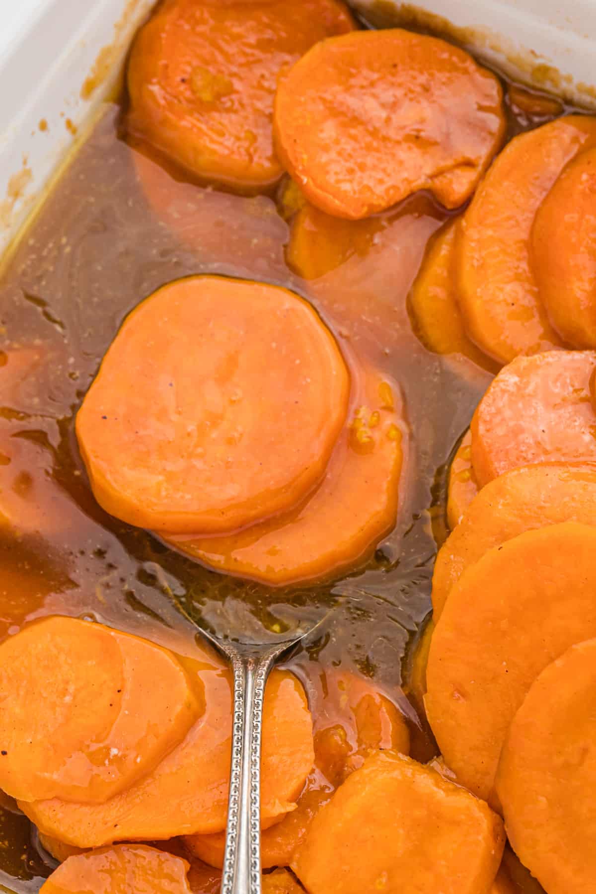 A silver spoon in a serving dish of candied yams in brown sugar syrup.