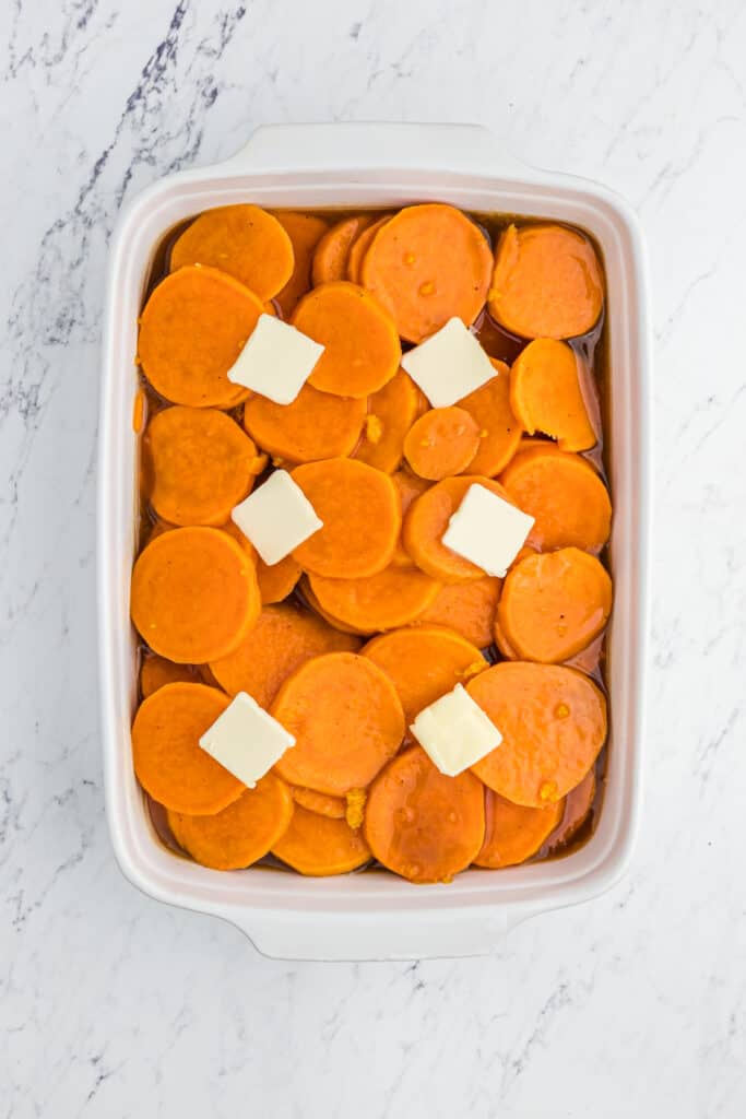 Sliced yams in a white baking dish topped with pats of butter on white countertop