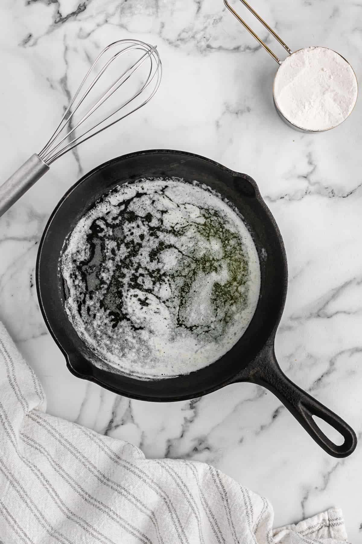 Melted butter in a cast iron skillet with a whisk and cup of flour nearby.