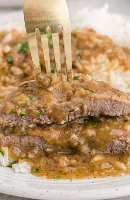 A fork showing a cut slice of smothered round steak on a white plate.