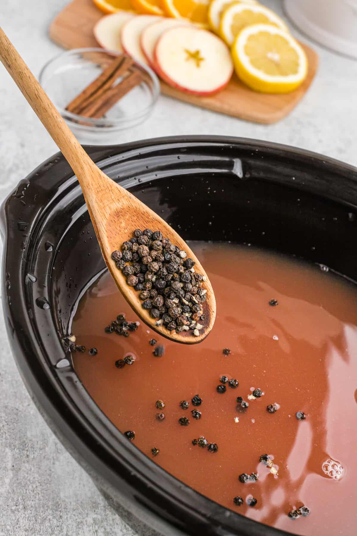 A wooden spoon adding whole peppercorns to a crockpot of wassail.
