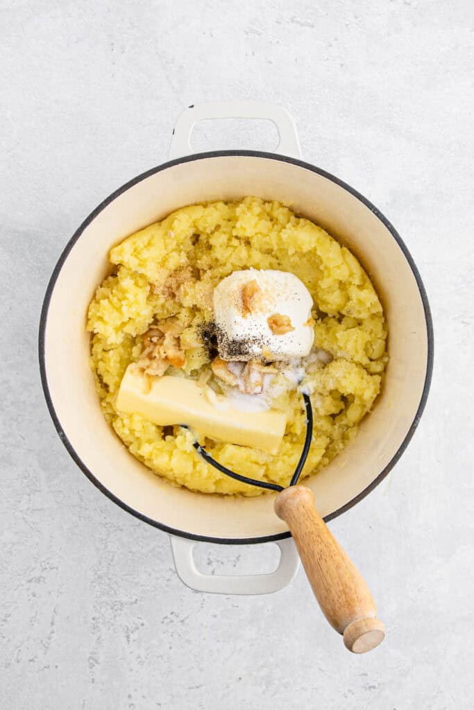Mashed potatoes with sour cream being mashed in a large pot with a masher on white countertop