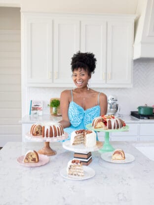 Jocelyn Delk Adams standing with three cakes on cake stands and smiling