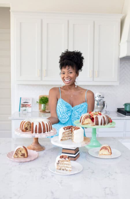 Jocelyn Delk Adams standing with three cakes on cake stands and smiling