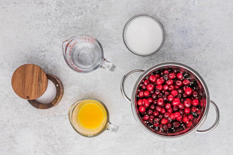 Ingredients to make cranberry sauce recipe in small bowls on a white surface.