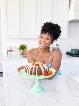 Jocelyn Delk Adams of Grandbaby Cakes holding a strawberry lemonade cake on a cake stand and smiling