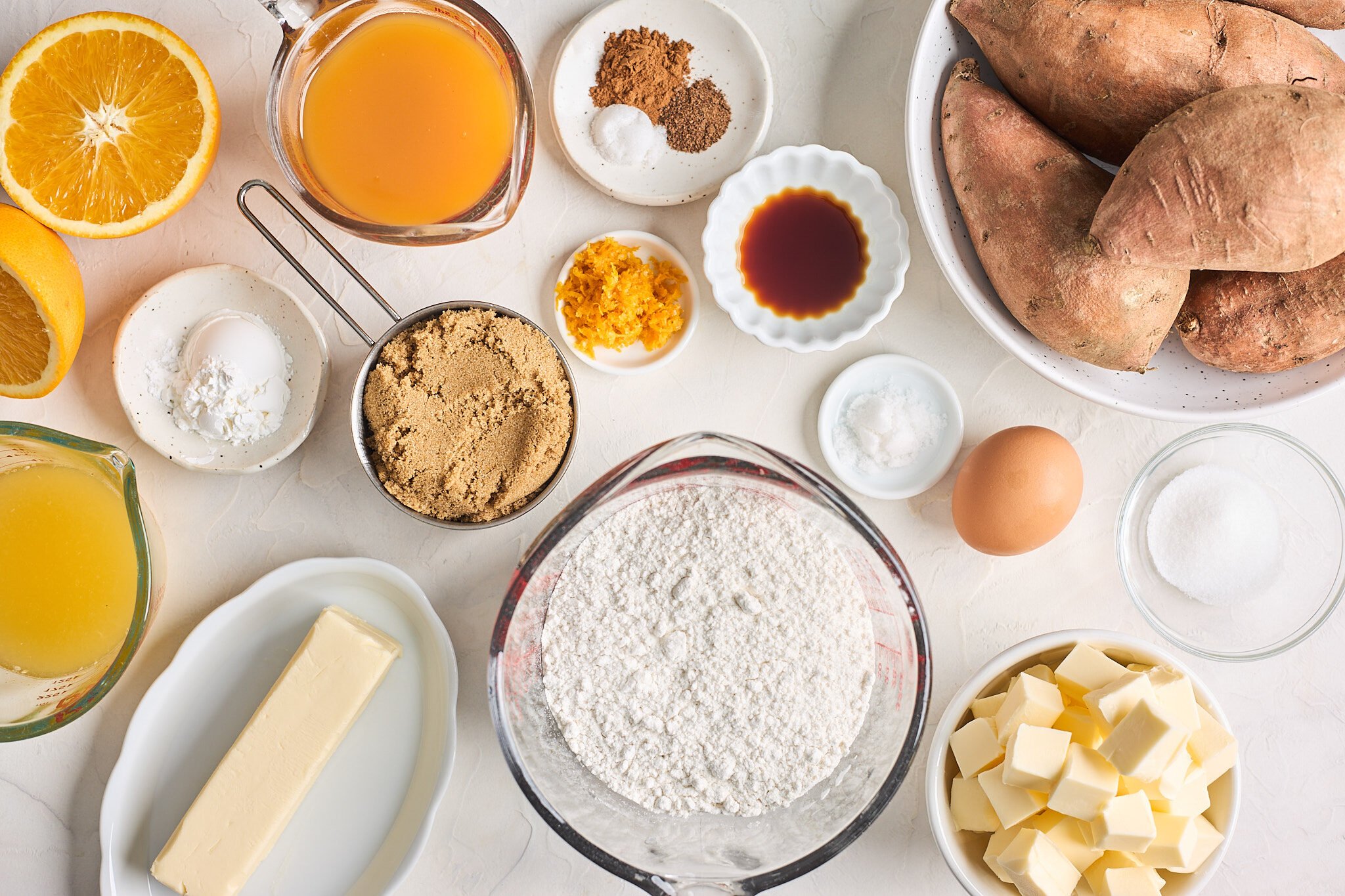 Sweet potatoes, butter, sugar, spices, vanilla and juices in clear and white bowls on white countertop