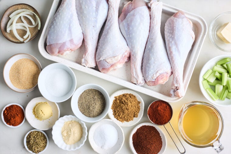 Turkey legs, several spices, chicken stock and butter in white bowls on white countertop