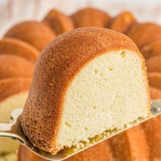 A slice of sour cream pound cake being removed from a full cake on a cake server in a white background