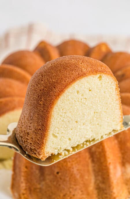 A slice of sour cream pound cake being removed from a full cake on a cake server in a white background