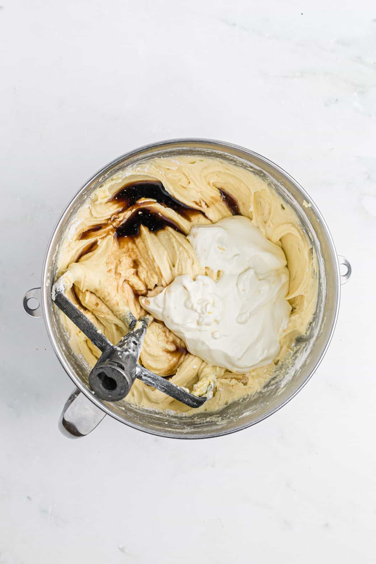 Sour cream and vanilla extract being added to a sour cream bundt cake batter in a stand mixer bowl on white countertop