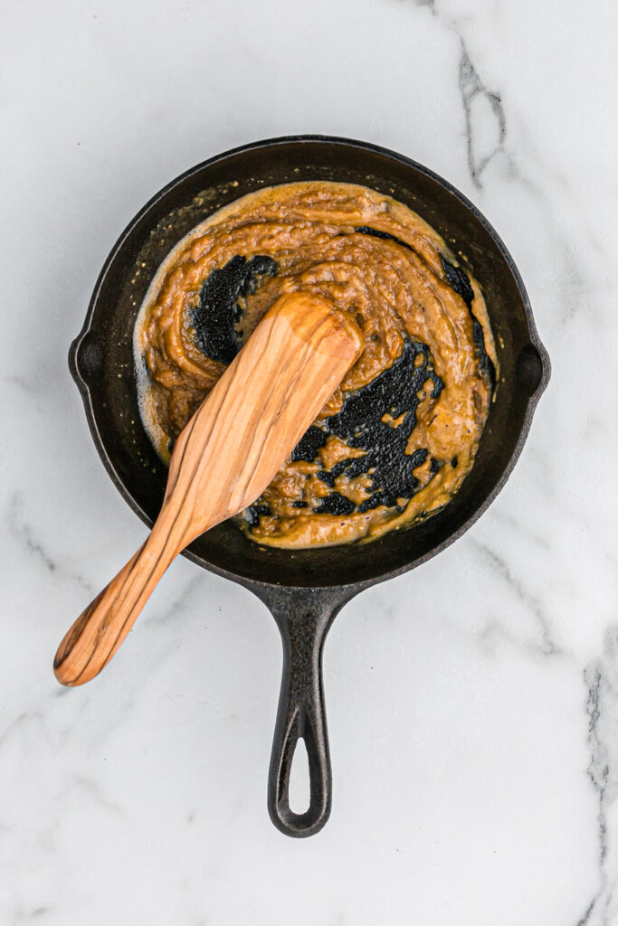 A wooden spoon stirring up a roux in a cast iron skillet on a white countertop