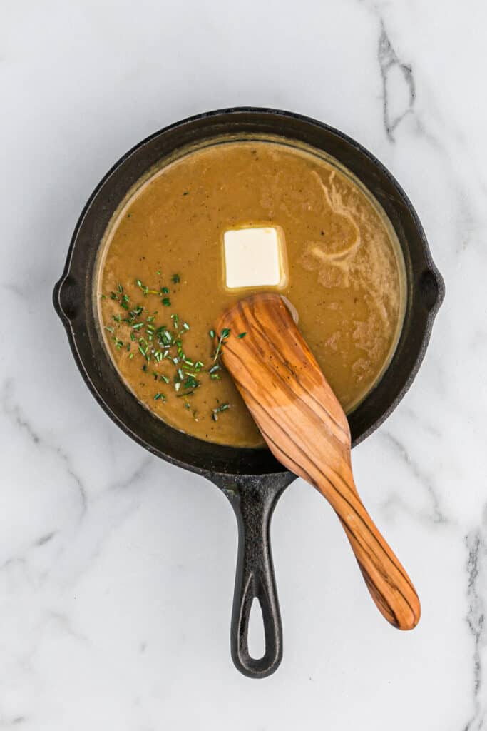 Butter and herbs being stirred into a saucepan of turkey gravy with drippings on a white countertop.