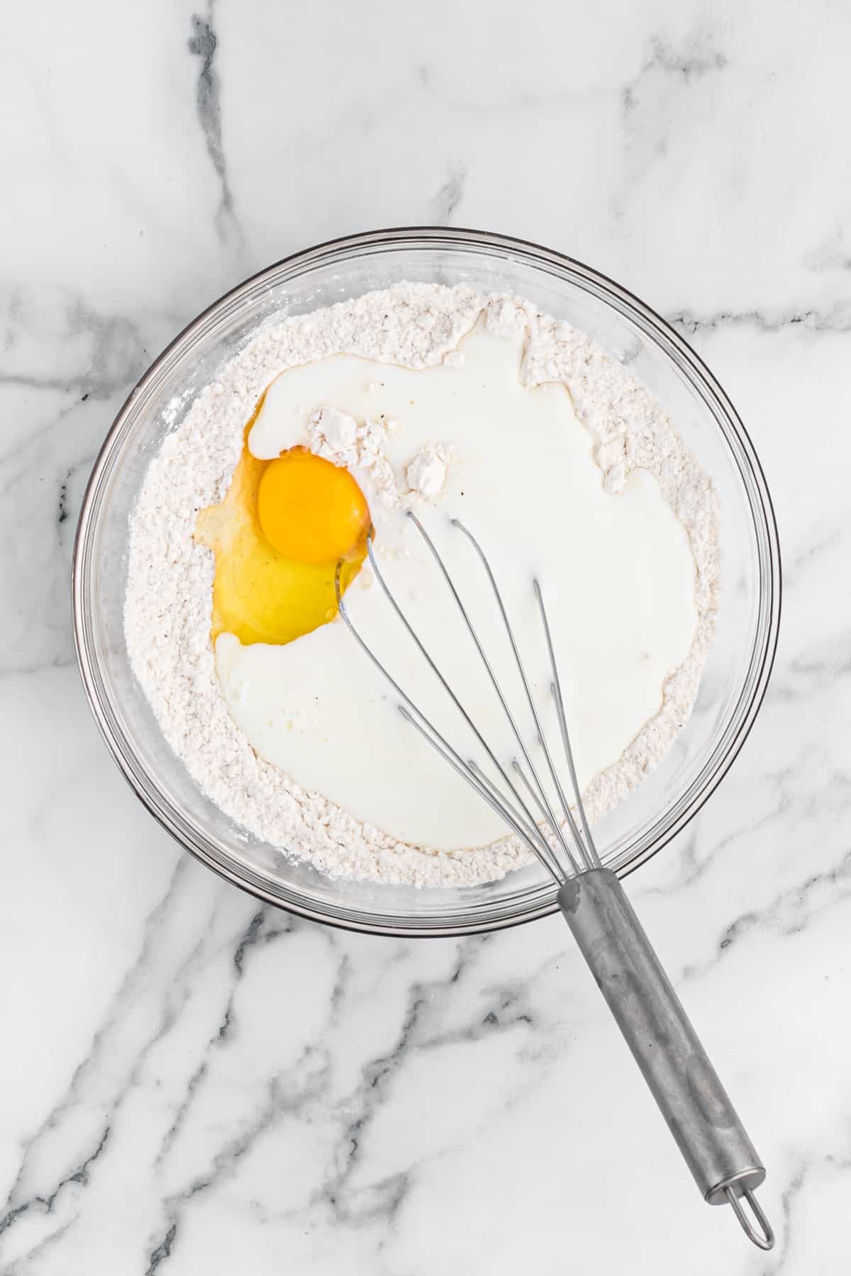 Egg and buttermilk being added to a flavored flour mixture in a glass bowl with a whisk on white countertop