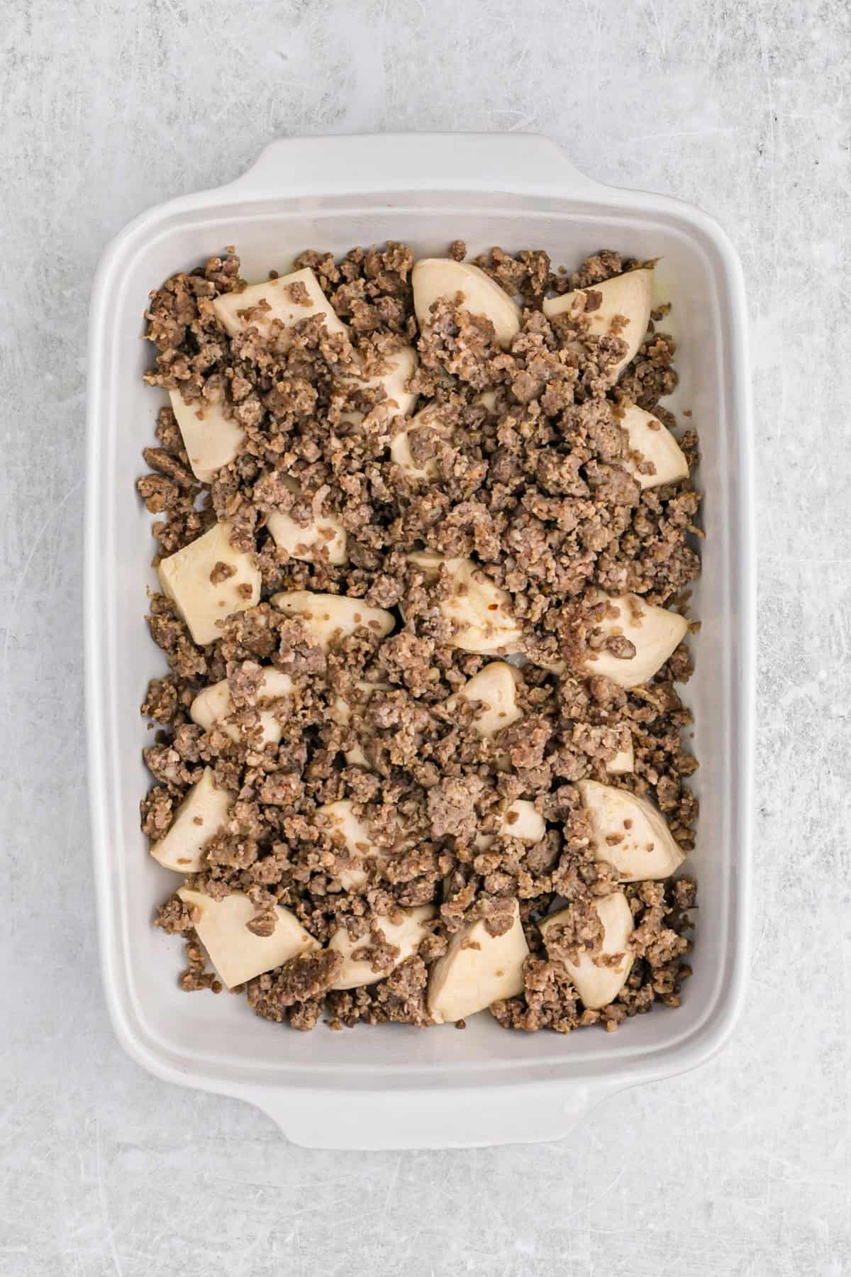 Crumbled cooked sausage layered on top of biscuit pieces in a white baking dish