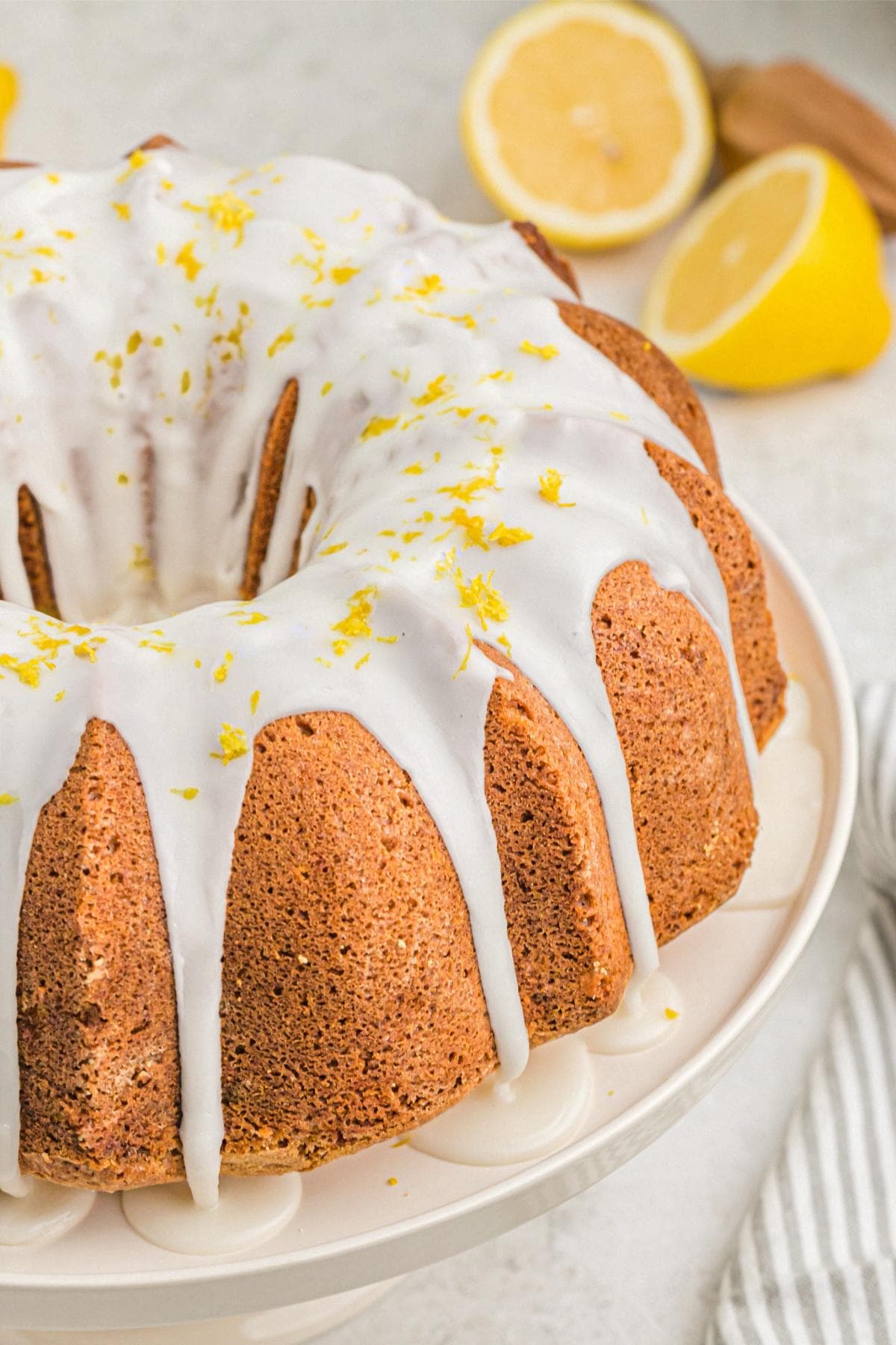 Glazed lemon bundt cake on a pedestal, finished with lemon zest and whole lemons nearby