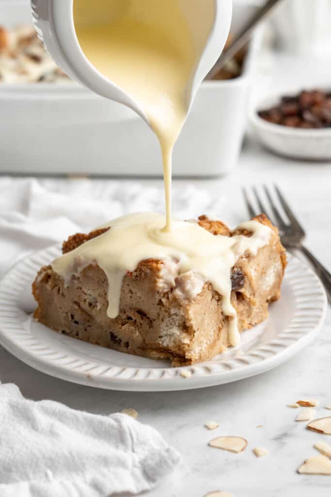 Creamy vanilla drizzle being poured over a slice of raisin bread pudding.