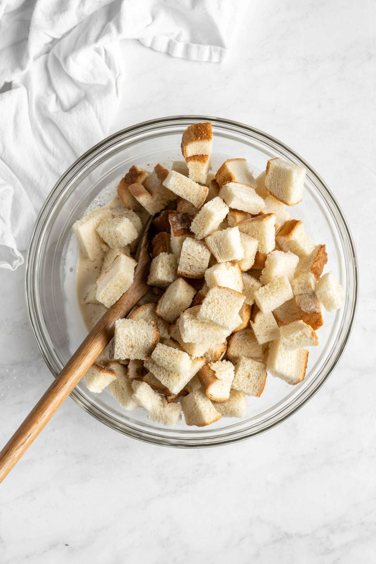 Dry bread cubes being added to custard in a glass bowl on white countertop