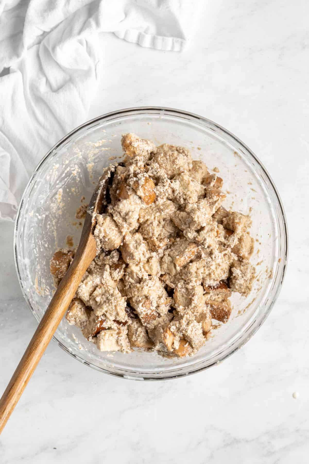 A wooden spoon coating bread cubes in bread pudding filling in a mixing bowl.