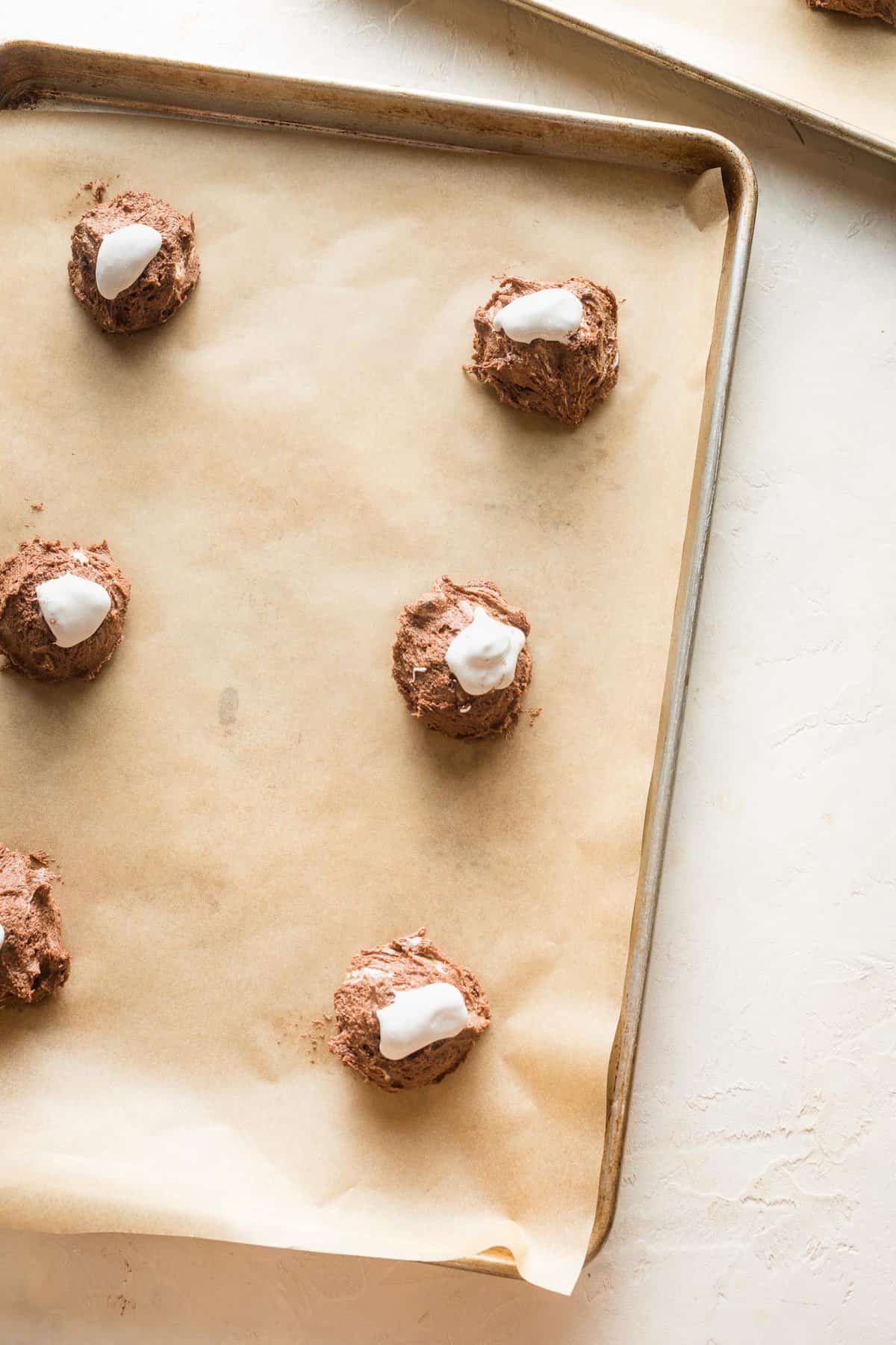 Hot cocoa cookie dough balls topped with marshmallow fluff on a parchment lined baking sheet.