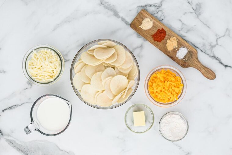 Cheese, sliced potatoes, spices, butter, flour and cream on white countertop