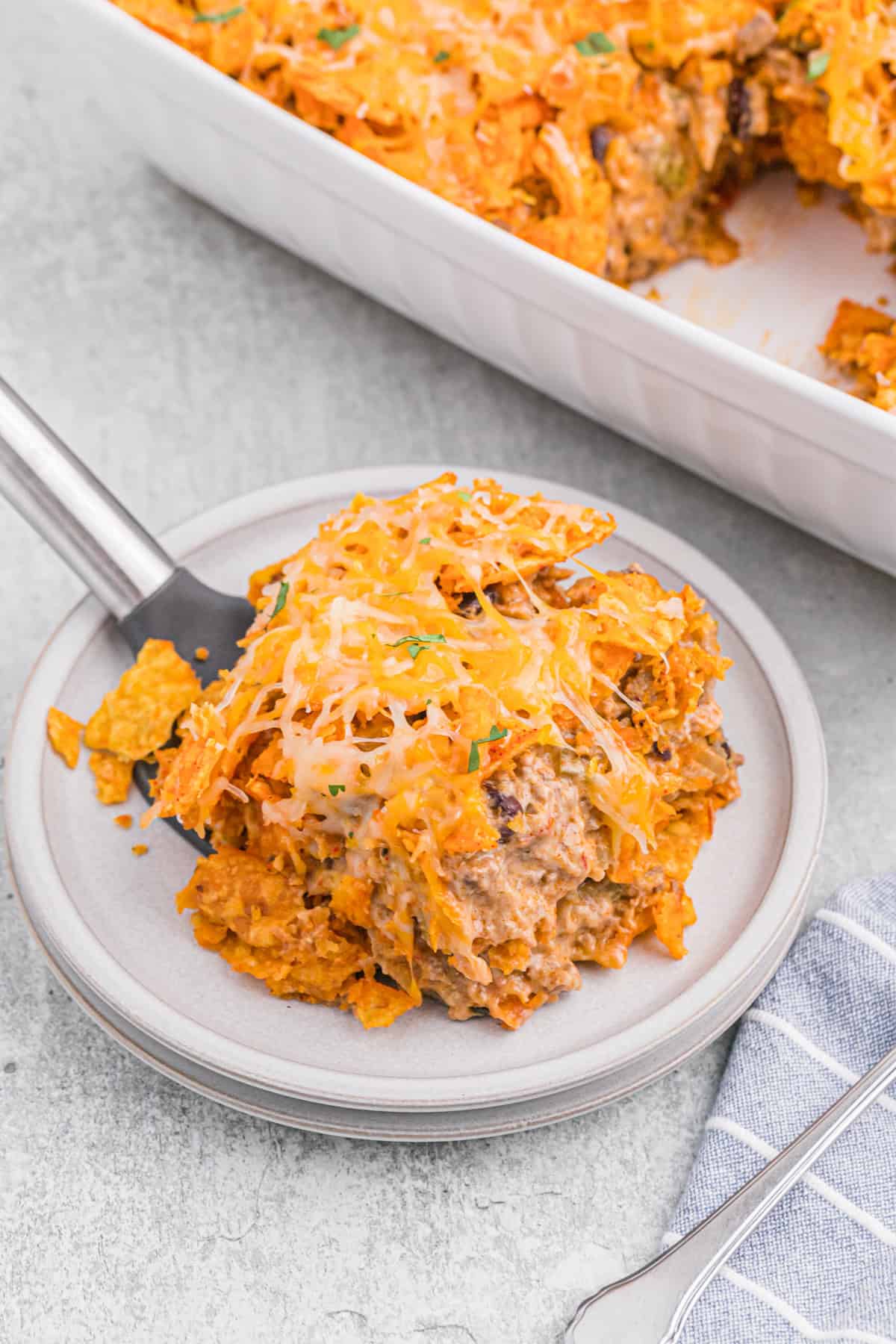 A plate with a serving of Dorito casserole, showing layers of chips, beef, and cheese, with the casserole dish in the background