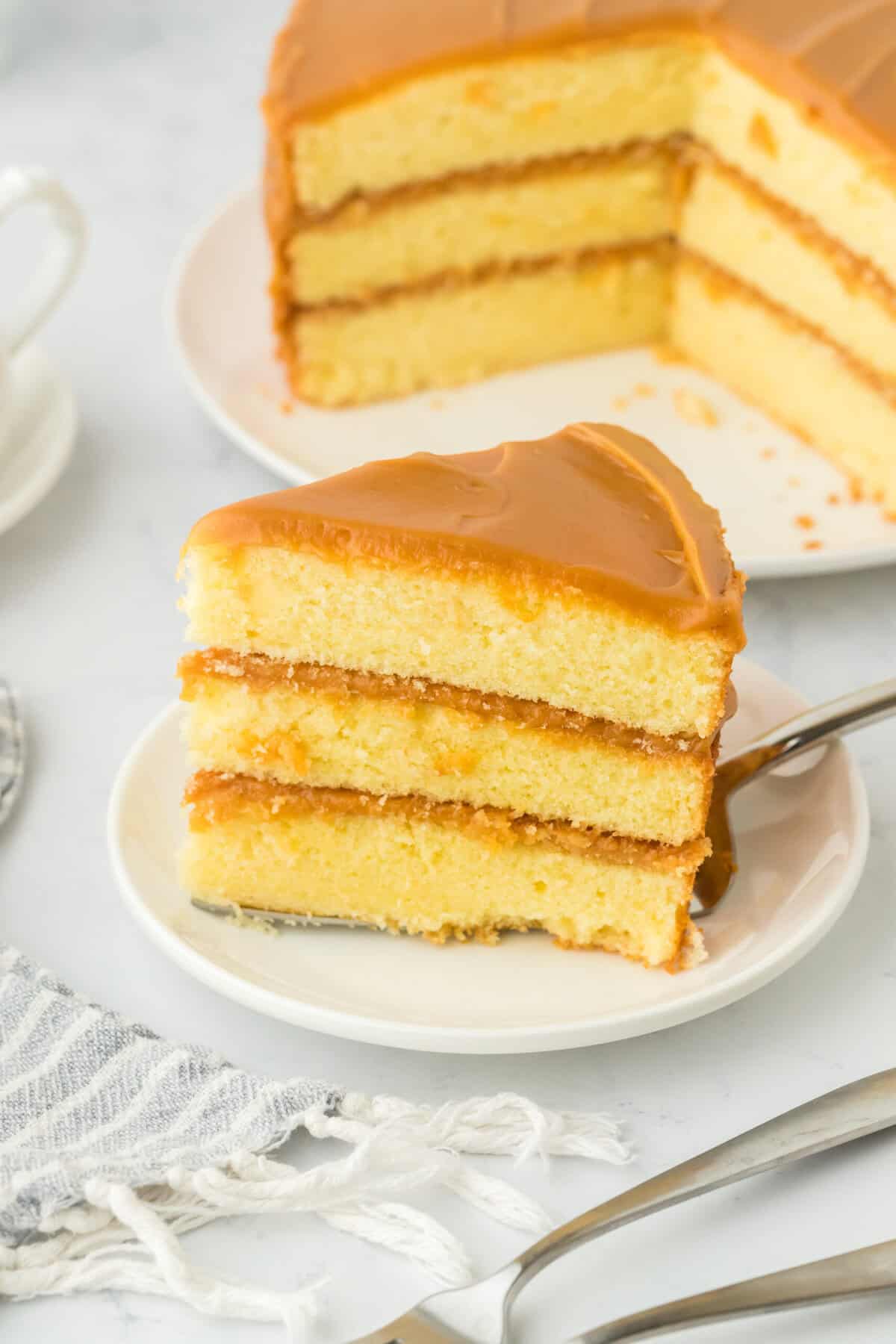 A slice of the best caramel cake recipe on a white plate under a cake server with full caramel cake in the white background