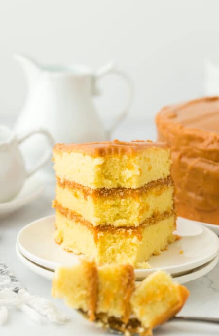 A slice of old fashioned caramel cake on a white plate with a fork full taken out on a white background