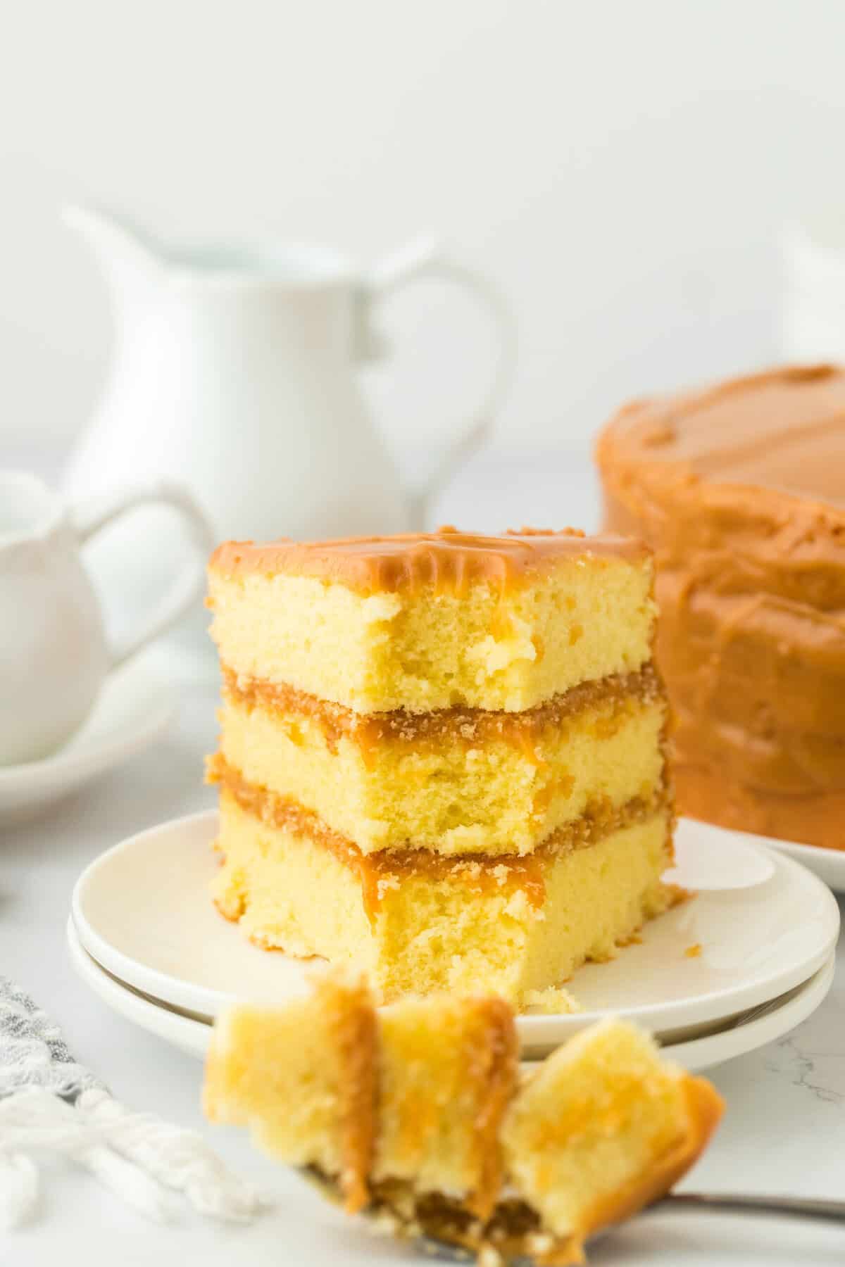 A slice of old fashioned caramel cake on a white plate with a fork full taken out on a white background