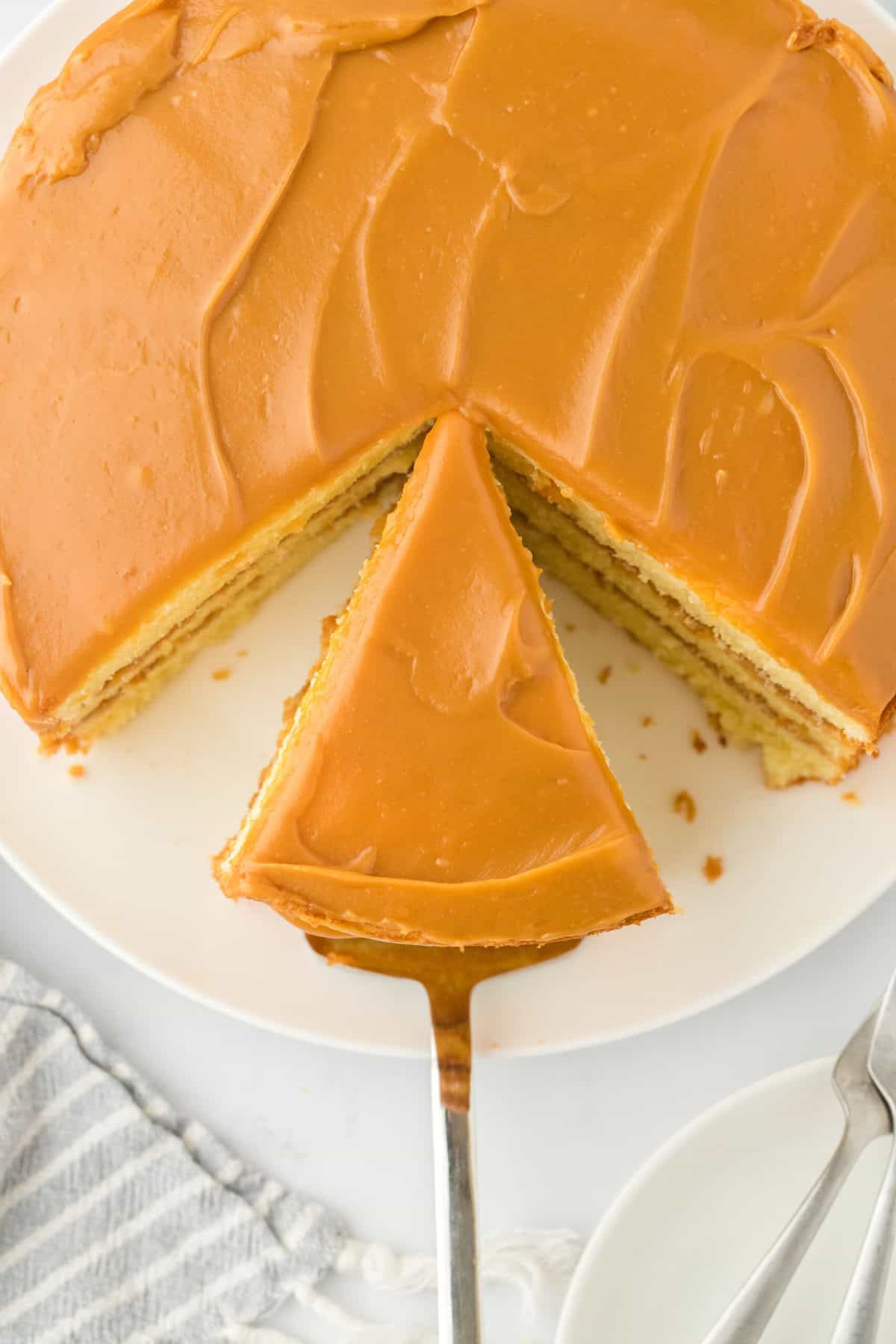 An overhead of a homemade caramel cake from scratch with a slice being taken out of the whole on a white cake stand on a server
