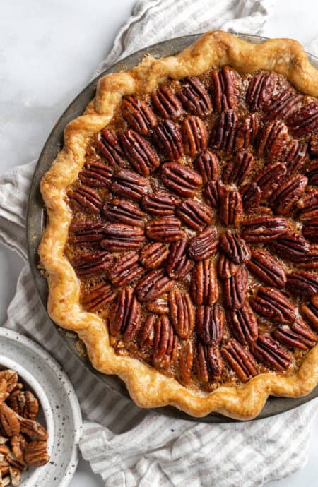 A perfect pecan pie recipe on a white background with a bowl of pecans nearby under a napkin